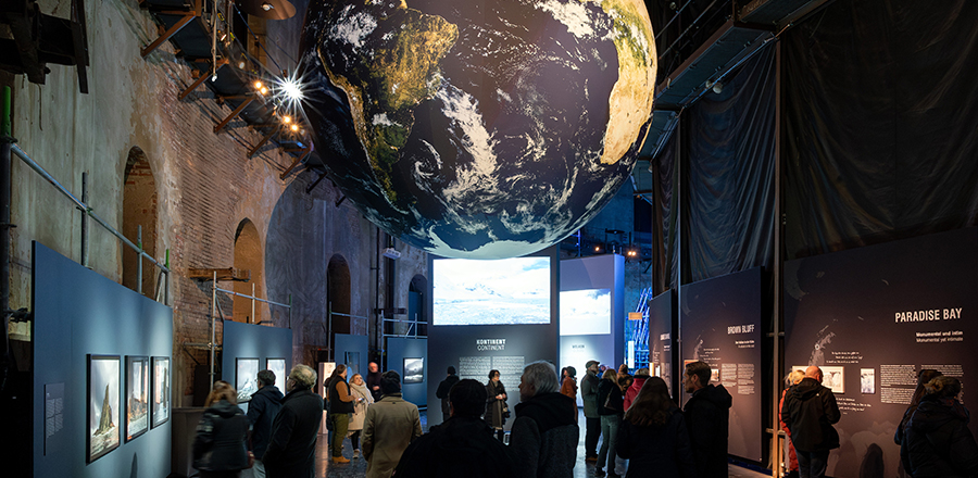 Zu sehen ist die ANTARKTIS-Ausstellung im Panometer Leipzig. In der Mitte hängt ein riesiger Globus im Raum sind Bilder zu sehen und viele Besucher