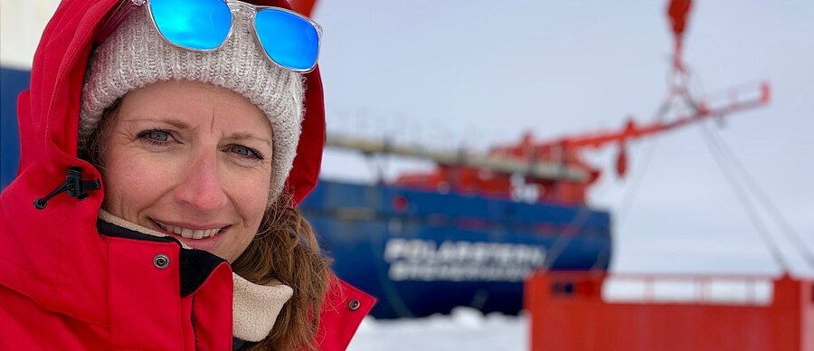 Forscherin in roter Winterbekleidung vor einem Forschungsschiff in der Antarktis